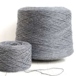 May include: Two skeins of grey yarn, one larger and one smaller, are shown on a white surface. The yarn is a medium weight and has a slightly textured appearance.