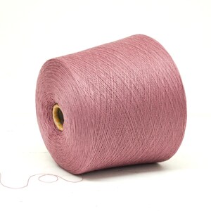 May include: A large spool of dusty rose yarn, ideal for knitting or crocheting projects. The yarn is tightly wound, showing fine strands, and sits on a white background. A strand of yarn extends from the spool.