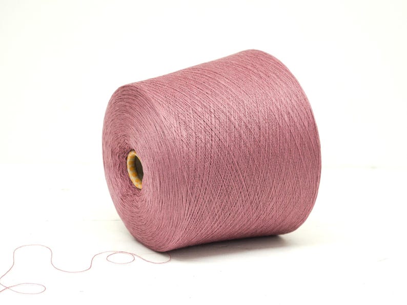 May include: A large spool of dusty rose yarn, ideal for knitting or crocheting projects. The yarn is tightly wound, showing fine strands, and sits on a white background. A strand of yarn extends from the spool.