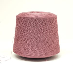 May include: A large spool of dusty rose-colored yarn. The yarn is tightly wound, forming a conical shape. A strand of yarn extends from the spool, suggesting it's ready for use in knitting or crocheting projects. The background is plain white.