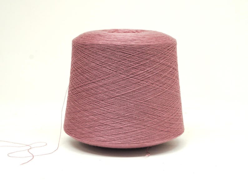 May include: A large spool of dusty rose-colored yarn. The yarn is tightly wound, forming a conical shape. A strand of yarn extends from the spool, suggesting it's ready for use in knitting or crocheting projects. The background is plain white.