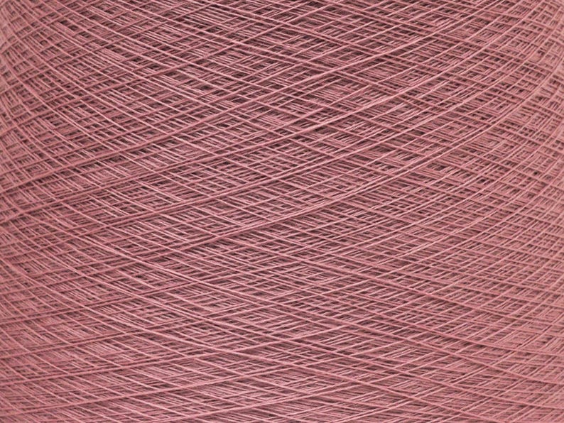 May include: Close-up of a large spool of dusty rose yarn. The yarn is tightly wound, creating a dense, textured surface. The threads are visible, forming a crisscross pattern. Ideal for knitting, crocheting, or other fiber arts projects.