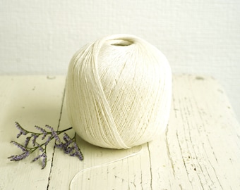 May include: A large ball of off-white yarn sits on a distressed, white wooden surface. A sprig of purple flowers rests near the yarn. The yarn is tightly wound, with a single strand extending from the ball. The background is a light, neutral color.