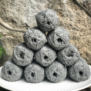Può includere: Un mucchio di dieci gomitoli di lana grigi disposti a forma di piramide su un piatto bianco. I gomitoli di lana sono fatti di un materiale morbido e testurizzato.