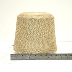 May include: A large cone of beige yarn sits on a white surface next to a metal ruler. The yarn is tightly wound, and the ruler shows measurements in inches. This yarn is suitable for various knitting and crochet projects.
