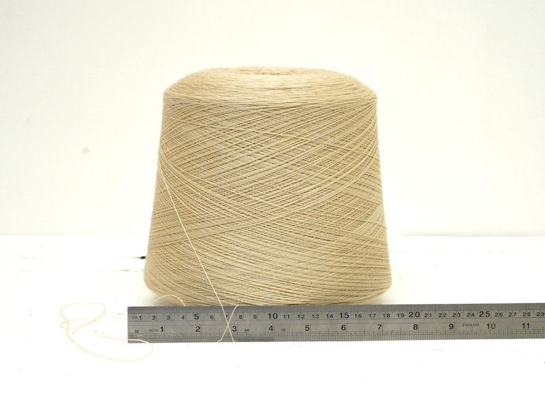 May include: A large cone of beige yarn sits on a white surface next to a metal ruler. The yarn is tightly wound, and the ruler shows measurements in inches. This yarn is suitable for various knitting and crochet projects.