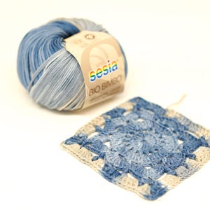 Może przedstawiać: Kłębek niebiesko-białej przędzy Sesia Bio Bimbo obok szydełkowego kwadratu. Przędza ma gradient jasnoniebieskiego i ciemnoniebieskiego. Szydełkowy kwadrat to wzór granny square w kolorze niebieskim i kremowym.