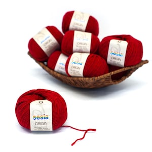 Può includere: Una collezione di gomitoli di lana rossi, alcuni in un cestino intrecciato e uno separato. Ogni gomitolo ha un'etichetta bianca con il testo "sesia ORIGIN". La lana è di un colore rosso vibrante e il cestino è marrone.
