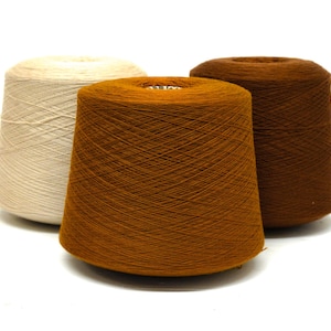 May include: Three spools of yarn in shades of cream, orange-brown, and dark brown. The yarn is tightly wound on conical spools, with the threads visible. The spools are arranged against a white background, highlighting the colors and textures.