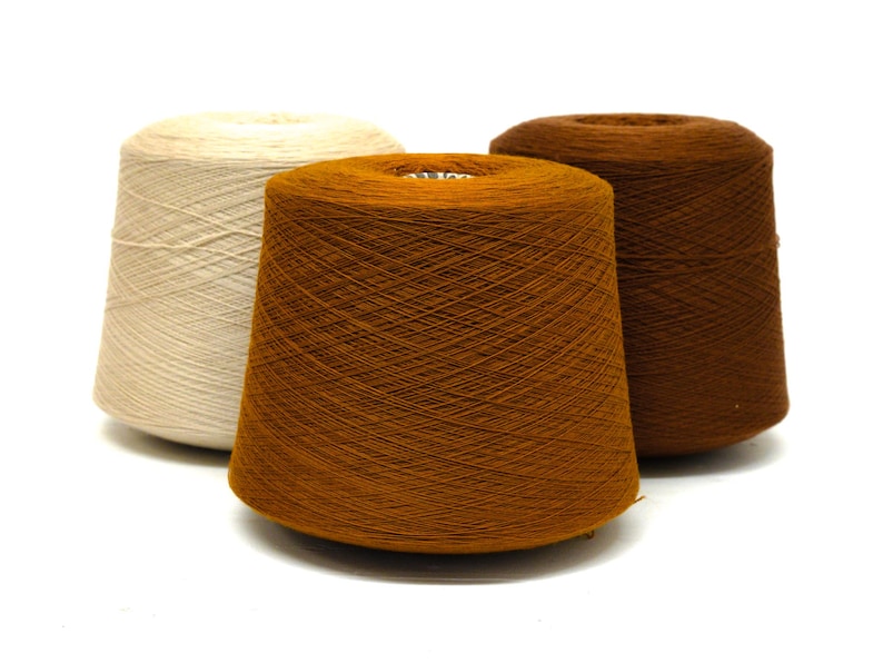 May include: Three spools of yarn in shades of cream, orange-brown, and dark brown. The yarn is tightly wound on conical spools, with the threads visible. The spools are arranged against a white background, highlighting the colors and textures.
