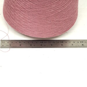 May include: A large cone of dusty pink yarn positioned behind a stainless steel ruler. The ruler shows the base of the yarn cone measures approximately 9.5 inches in width. A single loose strand of the pink yarn extends to the left side.