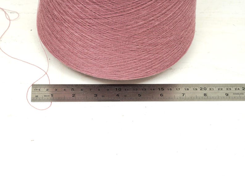 May include: A large cone of dusty pink yarn positioned behind a stainless steel ruler. The ruler shows the base of the yarn cone measures approximately 9.5 inches in width. A single loose strand of the pink yarn extends to the left side.