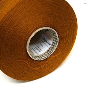 May include: A large spool of brown yarn, likely wool, is shown against a white background. The yarn is tightly wound, and the label on the core reads "Talleggo 1900". The yarn has a soft, textured appearance, ideal for knitting or crocheting projects.