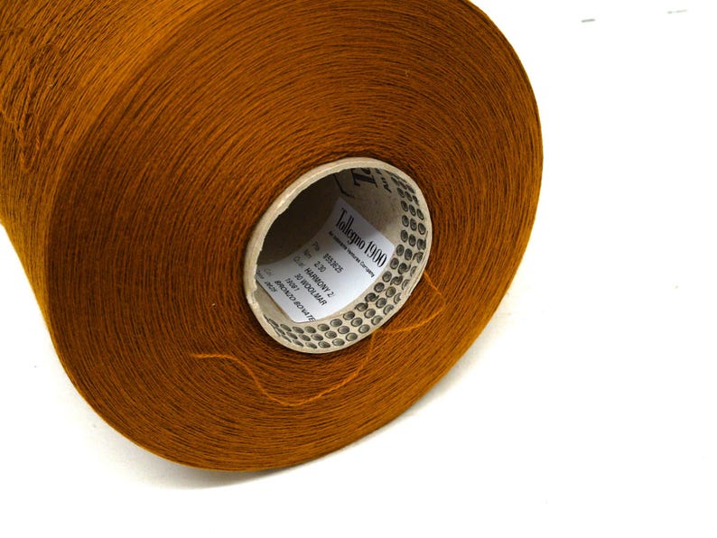 May include: A large spool of brown yarn, likely wool, is shown against a white background. The yarn is tightly wound, and the label on the core reads "Talleggo 1900". The yarn has a soft, textured appearance, ideal for knitting or crocheting projects.