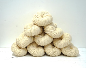 Puede incluir: Una pila piramidal de diez madejas de hilo color blanco roto. Cada madeja está enrollada en un paquete circular y retorcido, mostrando la textura suave y fibrosa del hilo. El color es un tono crema neutro y consistente en todas.