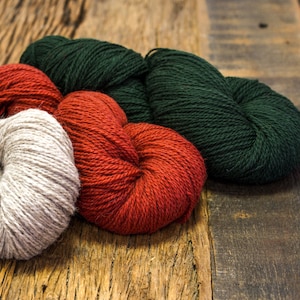 May include: Three skeins of yarn in different colors: a light beige, a dark red, and a dark green. The yarn is arranged on a wooden surface.