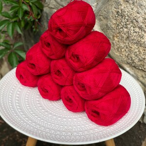 May include: A stack of eight red yarn balls on a white plate with a textured surface.