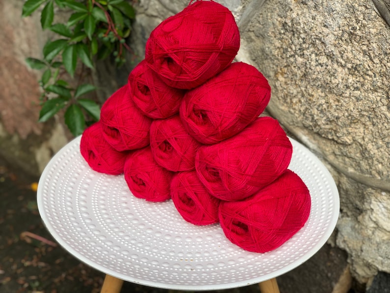 May include: A stack of eight red yarn balls on a white plate with a textured surface.