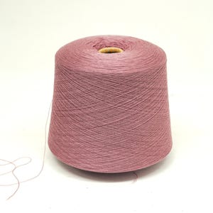 May include: A large spool of dusty rose-colored yarn. The yarn is tightly wound, forming a conical shape with a hollow core. A strand of yarn extends from the spool, suggesting it's ready for use in knitting or crocheting projects.