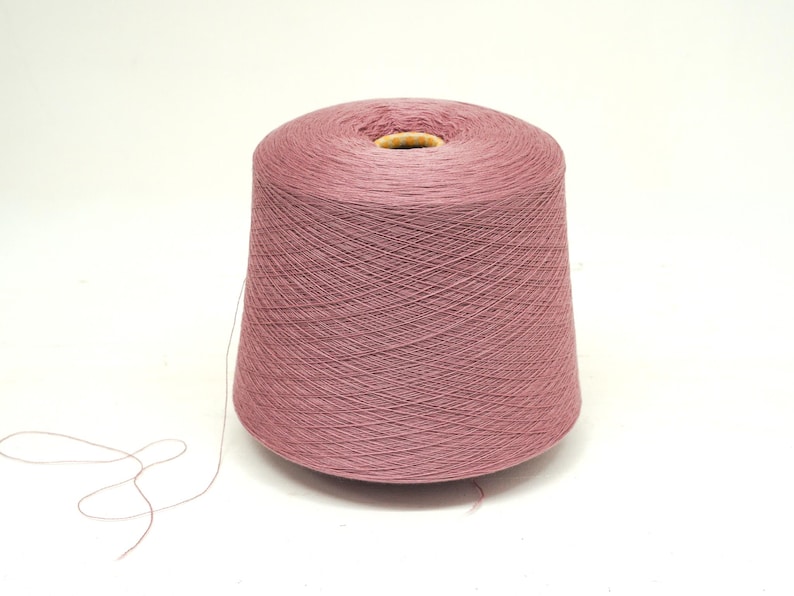 May include: A large spool of dusty rose-colored yarn. The yarn is tightly wound, forming a conical shape with a hollow core. A strand of yarn extends from the spool, suggesting it's ready for use in knitting or crocheting projects.