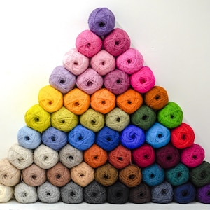 May include: A large pyramid arrangement of numerous yarn balls in a wide spectrum of colours including shades of purple, pink, yellow, orange, green, blue, red, brown, grey, black, and white, stacked neatly against a plain white background.