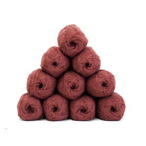 Può includere: Una pila piramidale di dieci gomitoli di lana rotondi di colore rosa antico scuro. Ogni gomitolo è avvolto con una texture in fibra visibile, creando un aspetto uniforme in tutta la collezione. Il filato è disposto in una formazione triangolare con un gomitolo in cima.