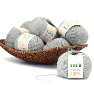 May include: A brown woven basket filled with several gray yarn balls. Each ball has a label that reads "sesia ORIGIN". One ball sits outside the basket. The yarn is a light gray color.