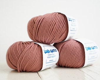May include: Three balls of Lana Gatto Maxi Soft yarn in a dusty rose color are stacked on a white surface. Each ball has a white label with blue text that reads "Lana Gatto Maxi Soft Made in Italy."