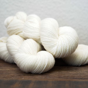 Peut inclure: Plusieurs écheveaux de fil de couleur ivoire, disposés sur une surface en bois foncé. Le fil est étroitement enroulé, avec un aspect doux et texturé. L'arrière-plan est gris clair neutre.
