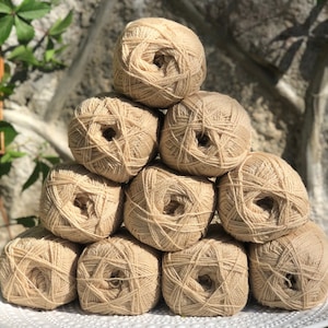 Op de afbeelding: Tien bollen beige garen gerangschikt in een piramidevorm op een wit bord. Het garen is zacht en pluizig, en de bollen zijn allemaal even groot.