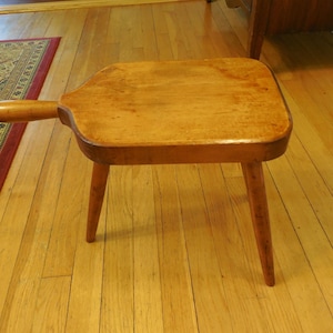 May include: A small wooden stool with a rectangular top featuring rounded corners and a single protruding handle on one side. The stool is supported by three tapered legs and has a smooth, light brown finish with visible wood grain patterns.