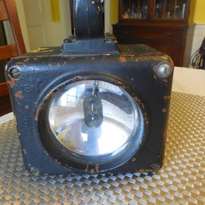 May include: A vintage black metal spotlight with a large, round glass lens. The spotlight has a handle on top and shows signs of wear, with chipped paint and rust. The light is sitting on a textured surface.
