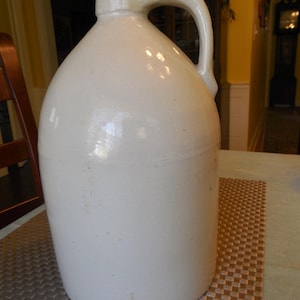 May include: A large, off-white stoneware jug with a rounded body and a handle. The jug has a wide base and a narrow neck, with a smooth, glossy finish. The jug is likely used for storing liquids and has a rustic, vintage aesthetic.