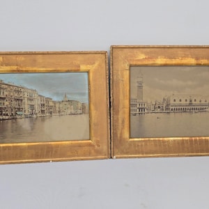 May include: Two framed prints of Venetian scenes. The frames are gold-colored with a distressed finish. One print depicts buildings along a canal with a blue sky, and the other shows a tower and buildings along the water.