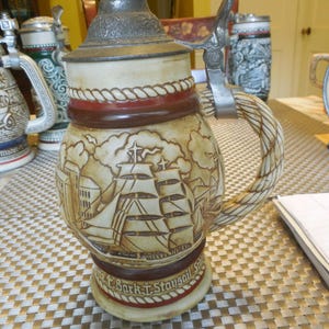May include: A vintage beer stein with a detailed nautical scene, featuring sailing ships and a coastal town. The beige ceramic stein has a metal lid and handle, with a rope-like design around the middle. The base reads "Bark & Staysail Schooner".