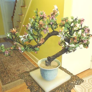 May include: A decorative bonsai tree with a brown trunk and branches, adorned with green leaves and pink, white, and orange flowers. The tree is potted in a light blue ceramic pot and sits on a decorative metal and marble table.