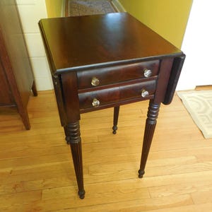 May include: A dark brown wooden side table featuring two stacked drawers with clear glass knobs. The table has a square top with drop-leaf sides and four slender, vertically fluted legs that taper toward the bottom, providing a stable base.