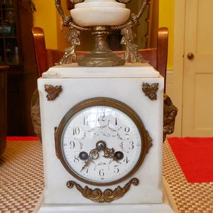 May include: Antique white marble clock with a round clock face and ornate gold-colored accents. The clock features a decorative top and base, with a detailed clock face showing the time. The clock is a vintage piece.