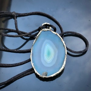 May include: A silver-rimmed pendant featuring a blue agate slice with a swirling pattern. The pendant is on a black cord.