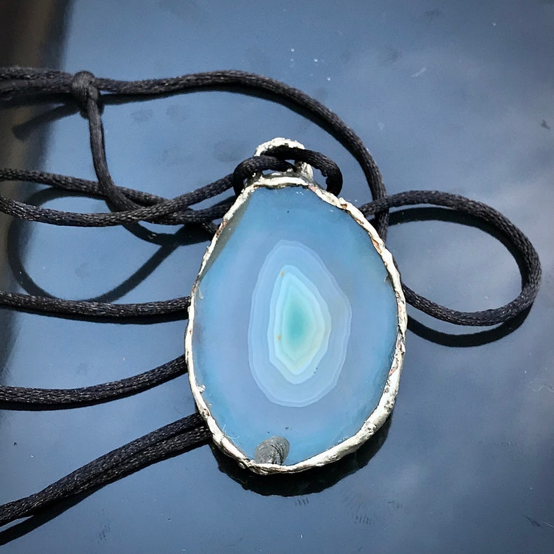 May include: A silver-rimmed pendant featuring a blue agate slice with a swirling pattern. The pendant is on a black cord.