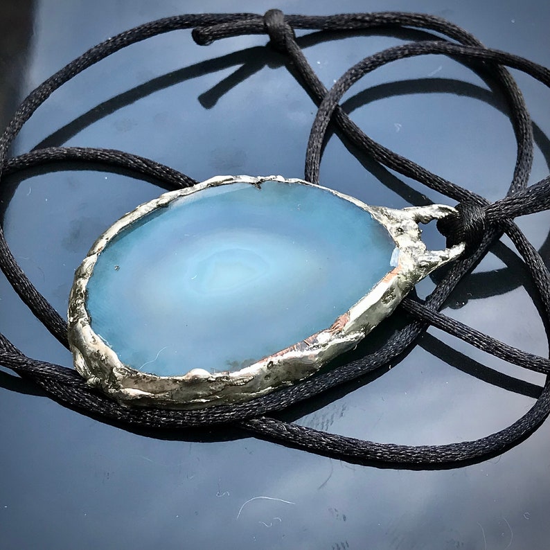 May include: A silver-toned pendant with a light blue agate stone. The pendant is on a black cord.