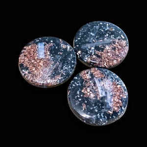 May include: Three round, clear resin objects with embedded copper and silver-colored metal pieces. The objects are on a black background, with the copper pieces appearing in a scattered pattern within the resin.