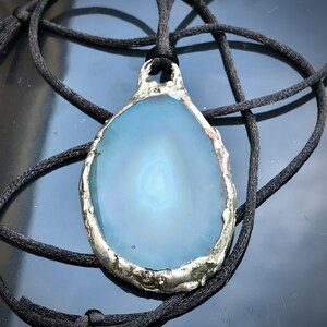 May include: A blue agate pendant with a silver-toned setting, hanging from a black cord.