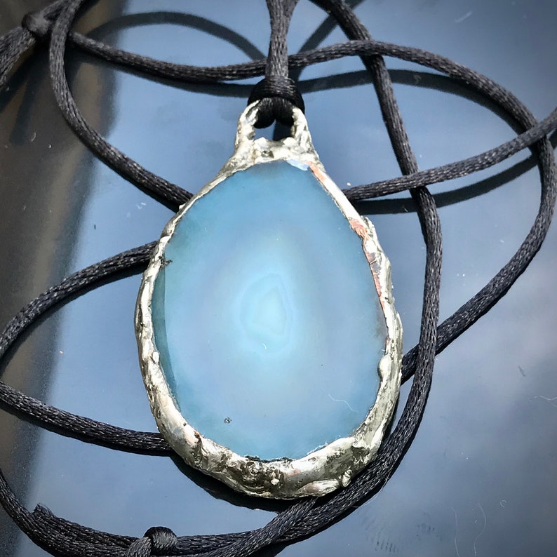 May include: A blue agate pendant with a silver-toned setting, hanging from a black cord.