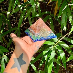 May include: A clear pyramid with a copper-colored base and a flower of life design. The pyramid is filled with green and blue crystals and has a copper spiral in the center.