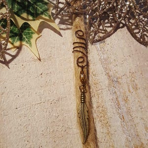 May include: A bronze-colored hair accessory featuring a feather charm and a spiral design. The accessory is attached to a strand of hair, with a gold-colored decorative element in the background. The overall aesthetic is bohemian and natural.