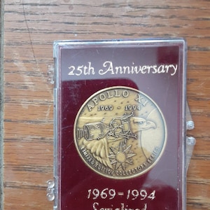 May include: A gold-colored coin in a red velvet case with a clear plastic cover. The coin features a depiction of the Apollo 11 spacecraft with the text "Apollo XI 1969-1994 Limited Edition Collectors Series" and "25th Anniversary 1969-1994 Serialized".