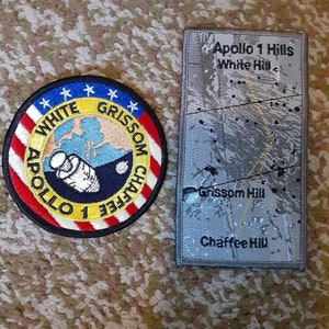 May include: Two embroidered patches, one round and one rectangular. The round patch is red, white, and blue with a yellow circle in the center. The text "APOLLO 1 GRISSOM CHAFFEE WHITE" is around the circle. The rectangular patch is gray with the text "Apollo 1 Hills White Hill Grissom Hill Chaffee Hill".