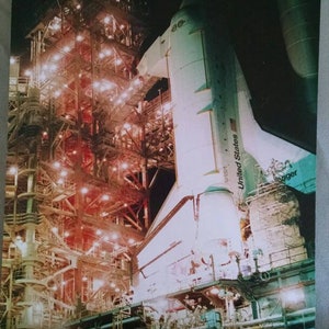 May include: A white space shuttle with the words "United States" on the side, is being prepared for launch. The shuttle is on a launch pad with a metal structure behind it. The image is titled "THE STS-8 FLIGHT COVER USPS/NASA".