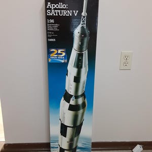 May include: A tall, rectangular Revell model kit box for the Apollo Saturn V rocket. The box features a graphic of the rocket against a space and earth background, with text including 1:96 scale, 114 cm, and a 25th anniversary 1969-1994 emblem.